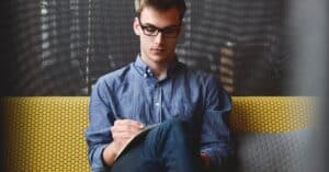 A young man in glasses writes in a notebook while sitting on a stylish couch indoors.